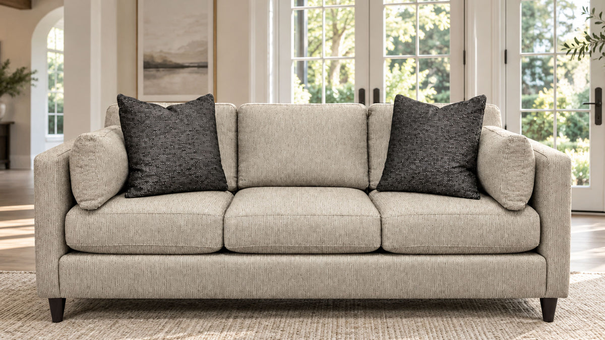 A three-seat upholstered sofa in a light beige textured fabric, styled with two dark gray textured throw pillows, set in a bright, modern living room with large windows.