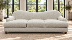 A modern three-seat sofa upholstered in a textured bouclé fabric in a light cream or off-white color, positioned in a bright living room with large black-framed windows and a woven rug.