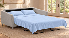 Un sofá cama tapizado en gris claro que se muestra en una habitación decorada con un juego de cama azul claro, que incluye dos almohadas y un edredón a juego, sobre una alfombra tejida junto a unas puertas acristaladas.
