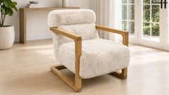 A modern armchair with a fluffy cream bouclé seat and back cushion, supported by a light natural wood frame, shown in a bright, minimalist room with light wood floors and large windows.