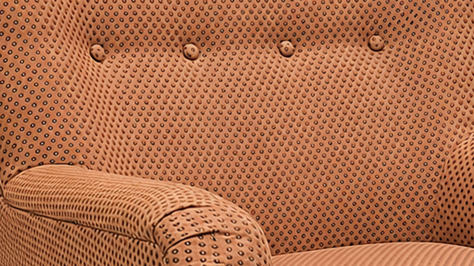 Close-up of a tufted upholstered armchair in a warm tan or camel fabric with a dense small-dot pattern and button tufting.