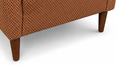 Close-up of the lower corner of an upholstered armchair with a warm brown fabric covered in small dark circular dots, showing part of the seat, side seam, and wooden legs on a white background.