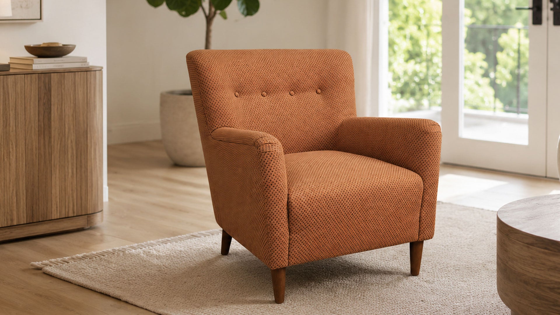 An orange textured armchair with button tufting on the backrest and tapered wooden legs, set in a bright, modern living room with light wood floors, a beige rug, and a glass door in the background.