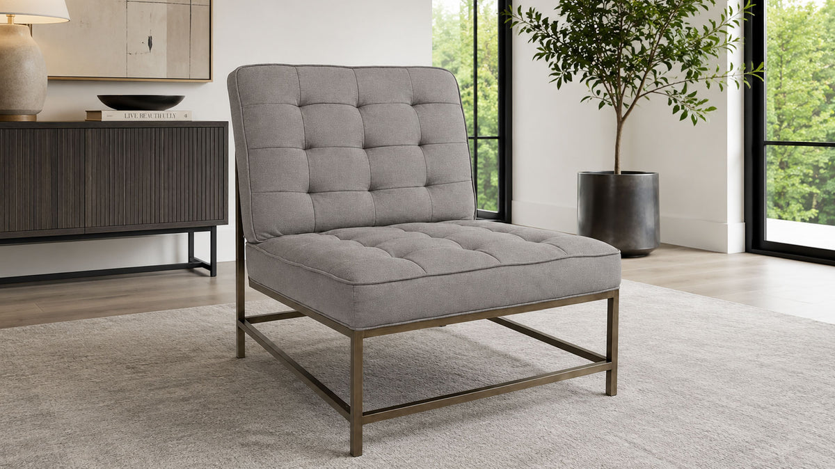 A modern tufted lounge chair with a light gray upholstered seat and backrest on a slim bronze metal frame, shown in a bright contemporary living room setting.
