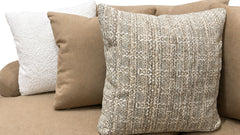 Three decorative throw pillows on a beige sofa: a large front pillow in textured woven beige and cream, a medium tan pillow, and a white bouclé pillow behind.