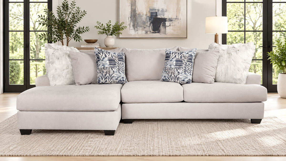Un sofá modular de color beige claro con una chaise longue orientada hacia la izquierda, decorado con varios cojines decorativos en un salón luminoso y moderno.