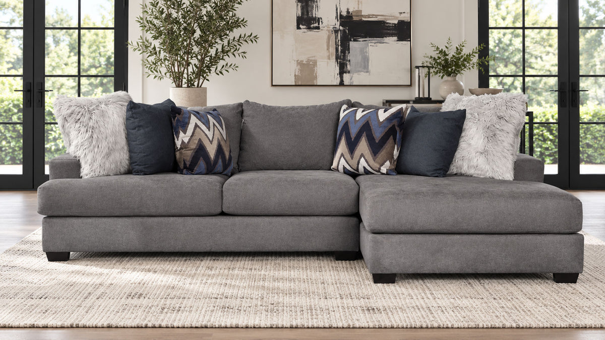 Un sofá modular de tela gris con una chaise longue orientada hacia la derecha, decorado con varios cojines decorativos en un salón luminoso y moderno.