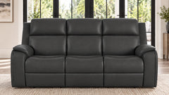 A three-seat black leather reclining sofa with padded back cushions and wide armrests, shown in a bright living room setting with large windows and a woven rug.