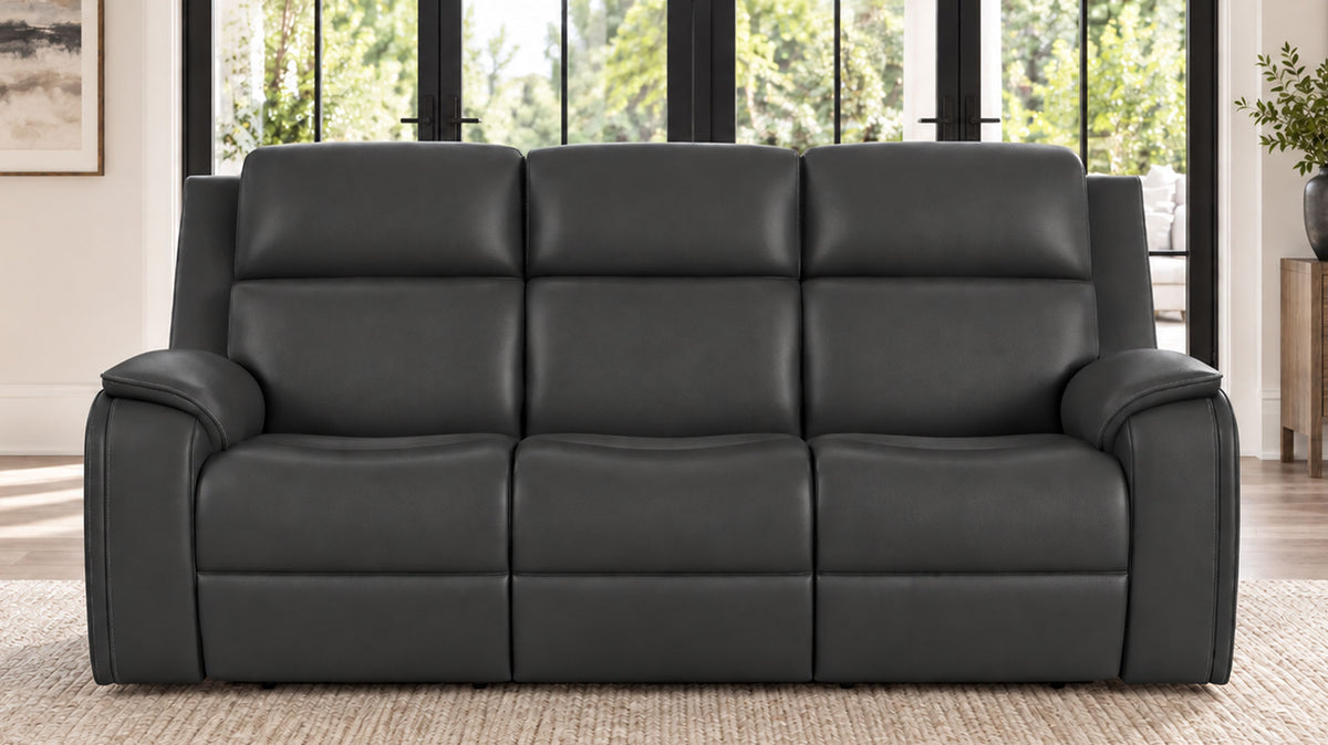 A three-seat black leather reclining sofa with padded back cushions and wide armrests, shown in a bright living room setting with large windows and a woven rug.