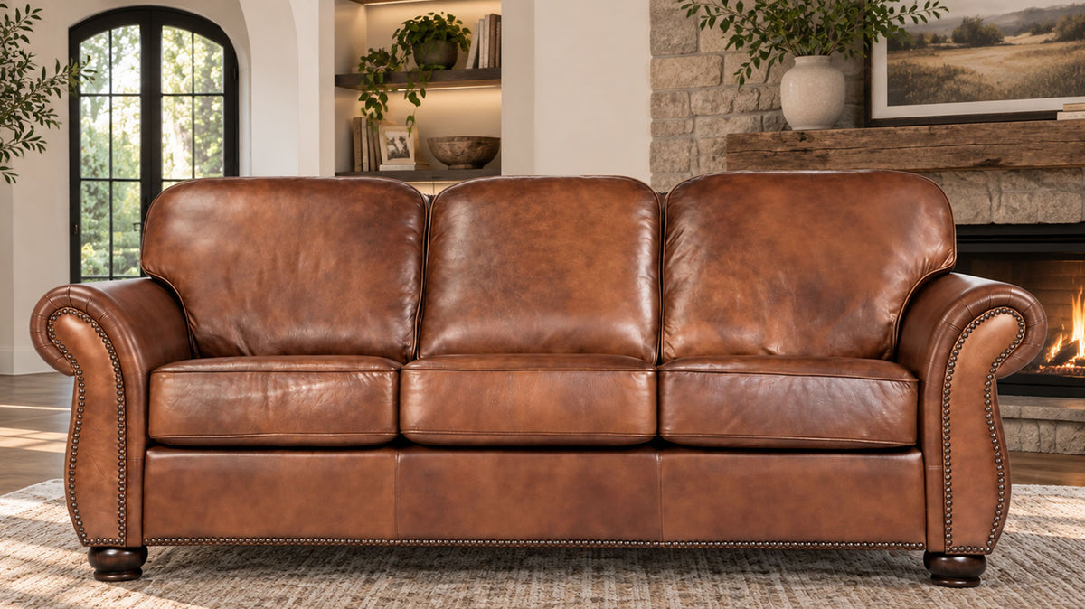 A three-seat brown leather sofa with rolled arms and nailhead trim, shown in a styled living room setting with a fireplace and window in the background.