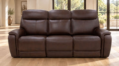 A three-seat reclining leather sofa in dark brown, positioned in a bright living room with light wood floors and large windows.