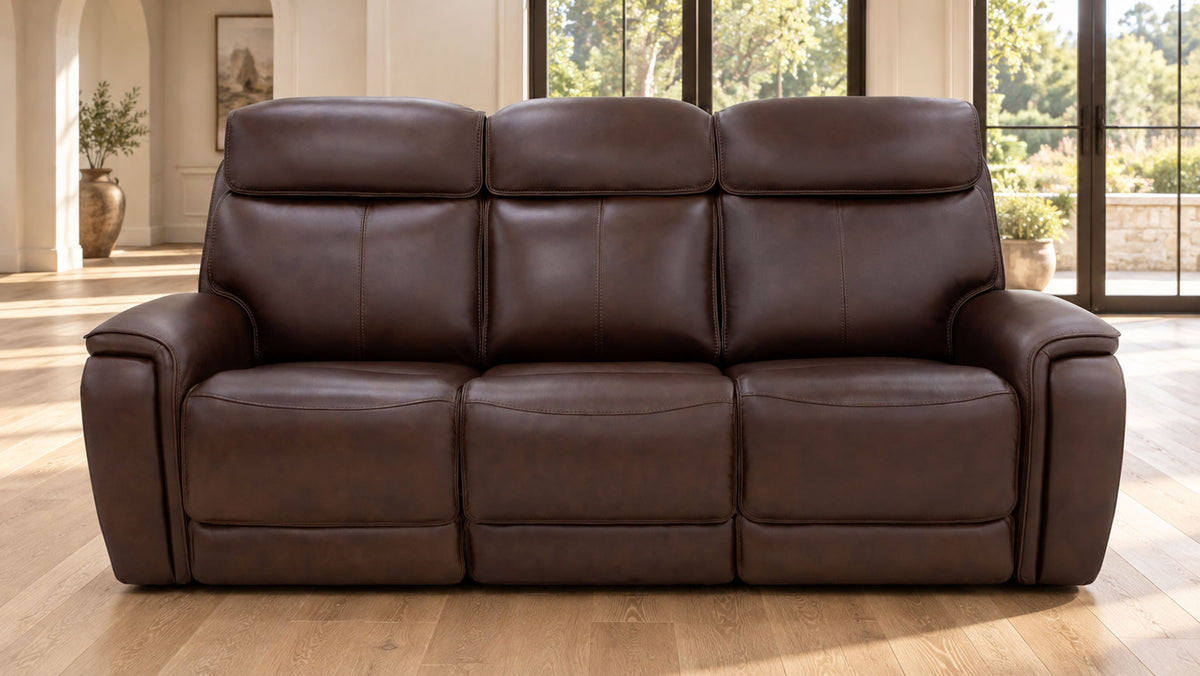 A three-seat reclining leather sofa in dark brown, positioned in a bright living room with light wood floors and large windows.