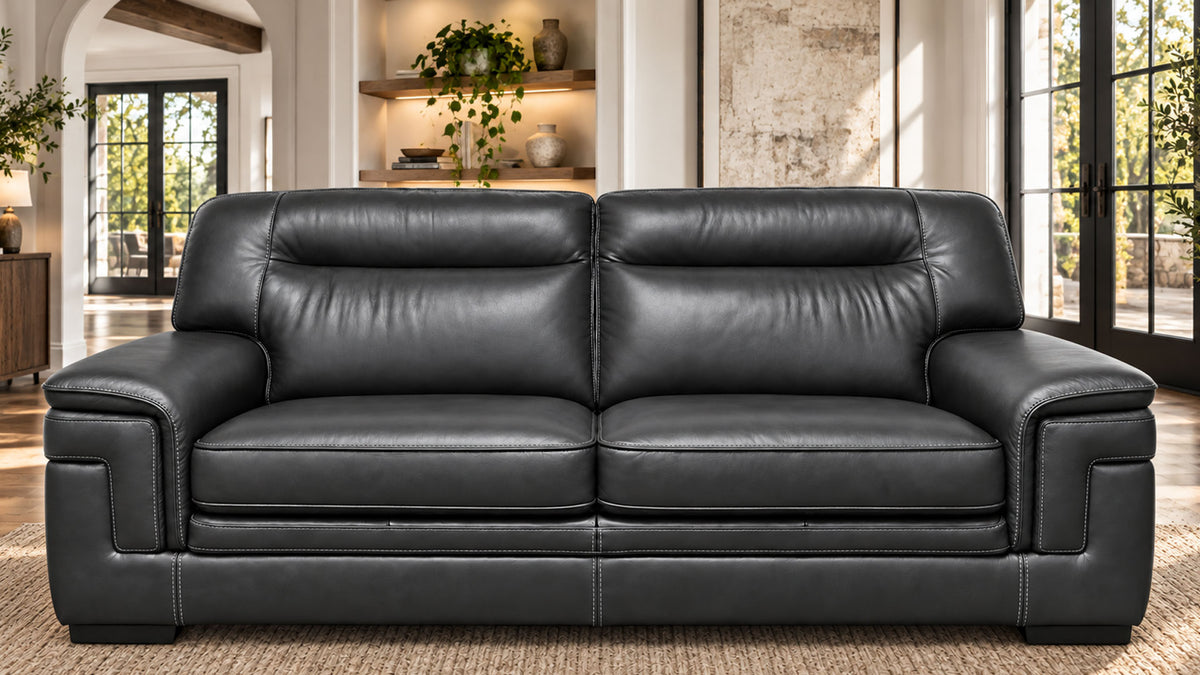 A black leather loveseat sofa with deep seat cushions and wide armrests, shown in a bright, modern living room with large windows and built-in shelving.