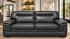 A black leather loveseat sofa with deep seat cushions and wide armrests, shown in a bright, modern living room with large windows and built-in shelving.