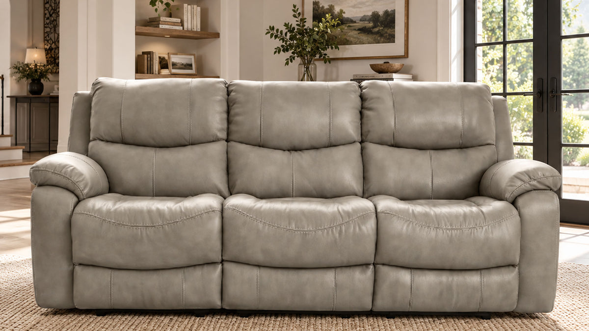 A three-seat reclining leather sofa in a light gray color, positioned in a modern living room setting with a woven rug, shelving, and large windows.