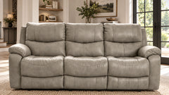 A three-seat reclining leather sofa in a light gray color, positioned in a modern living room setting with a woven rug, shelving, and large windows.