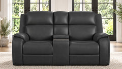 A black leather reclining loveseat sofa with a center console armrest, shown in a bright living room setting with large windows and plants.
