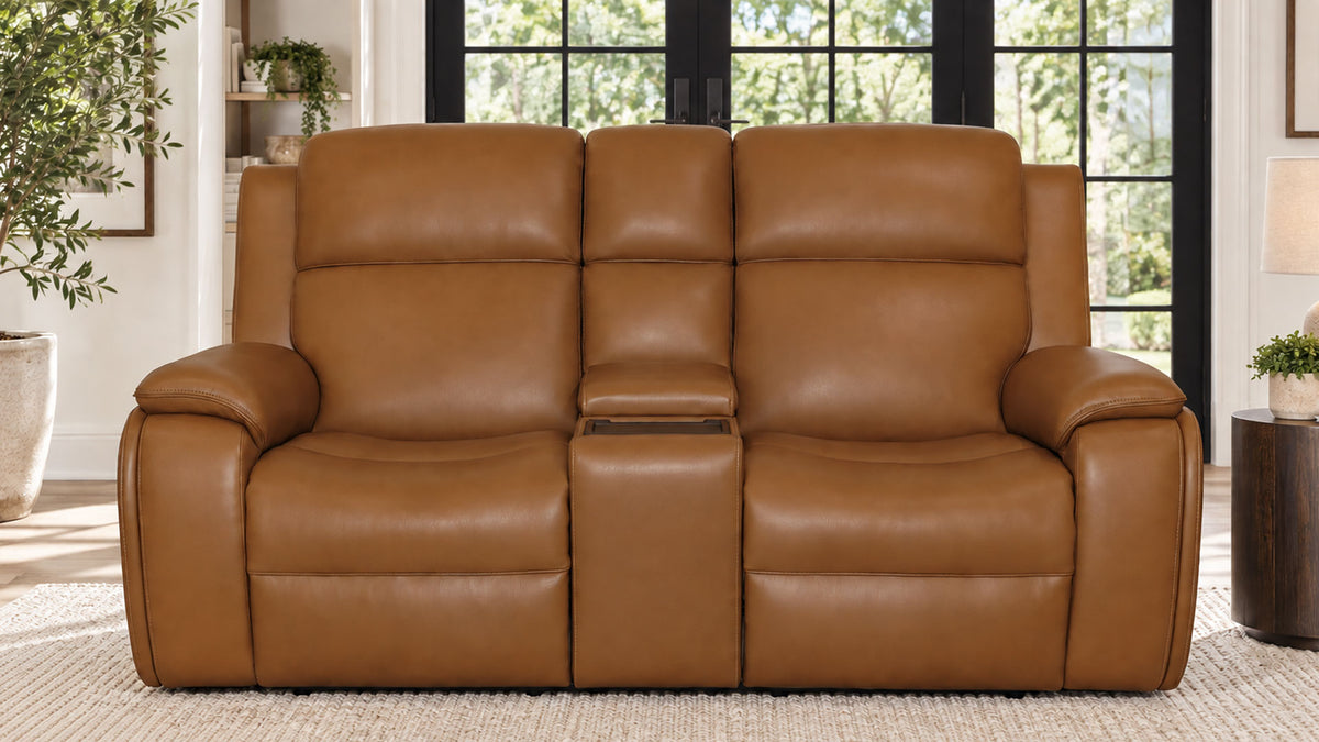 A three-seat power reclining loveseat in smooth tan leather, positioned in a bright living room setting with large black-framed windows and plants in the background.