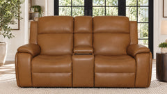 A three-seat power reclining loveseat in smooth tan leather, positioned in a bright living room setting with large black-framed windows and plants in the background.