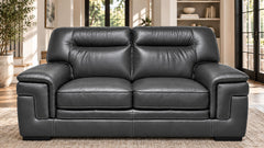 A black leather loveseat sofa with two seat cushions and padded backrests, shown in a bright living room setting with windows, shelving, and a rug.