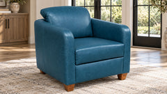 Un sillón azul de imitación de piel con patas de madera, situado sobre una alfombra beige con textura en un salón luminoso con grandes ventanales y un armario de madera al fondo.