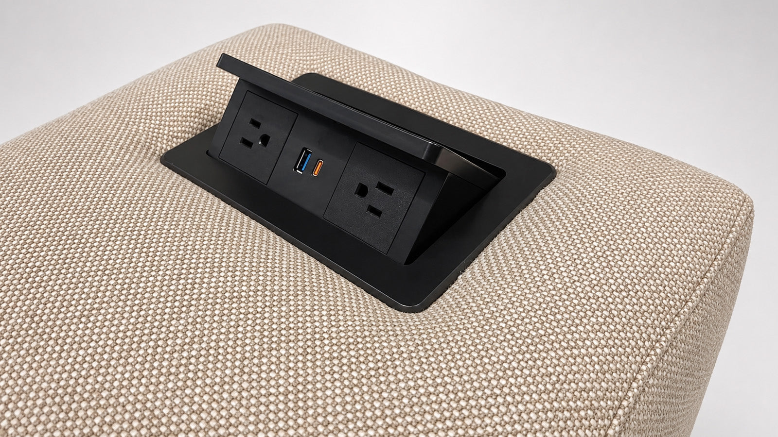 Close-up of a beige woven upholstered surface with a black recessed power outlet panel featuring two outlets and a USB-C port, shown on a light gray studio background.