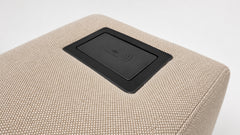 Close-up of a beige woven-texture ottoman or sofa cushion with a black rectangular wireless charging pad inset on top, shown against a clean white background.