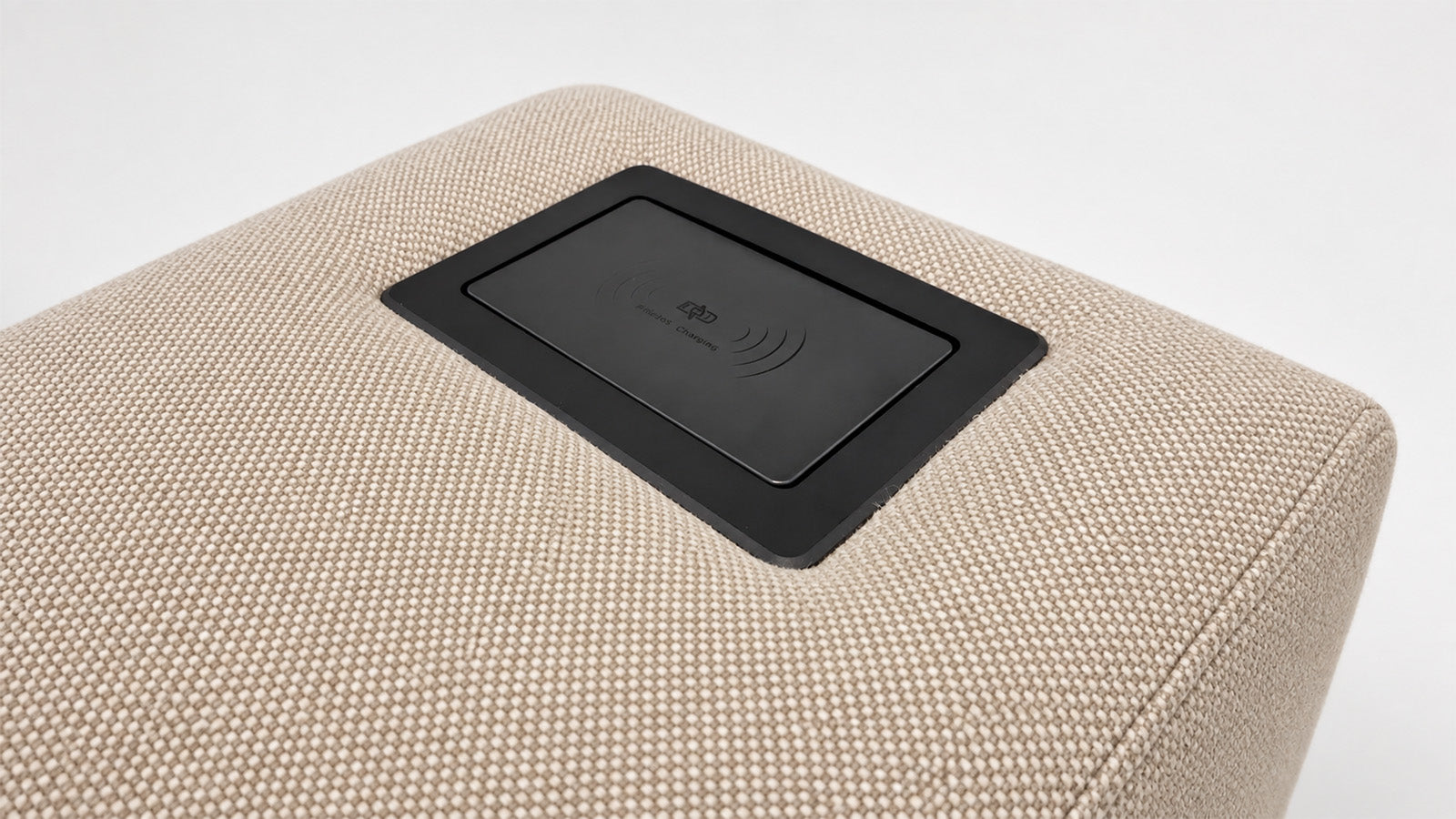 Close-up of a beige woven-texture ottoman or sofa cushion with a black rectangular wireless charging pad inset on top, shown against a clean white background.