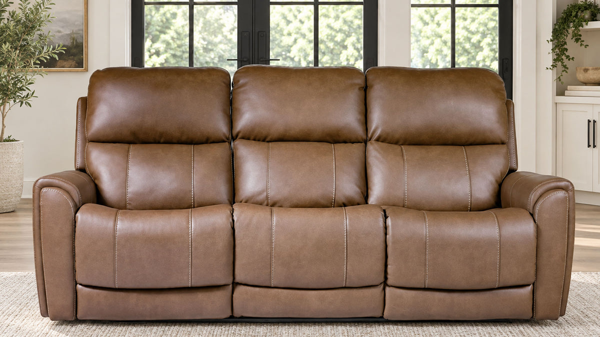 A three-seat power reclining loveseat sofa upholstered in brown faux leather, positioned in a bright living room setting with windows and plants in the background.