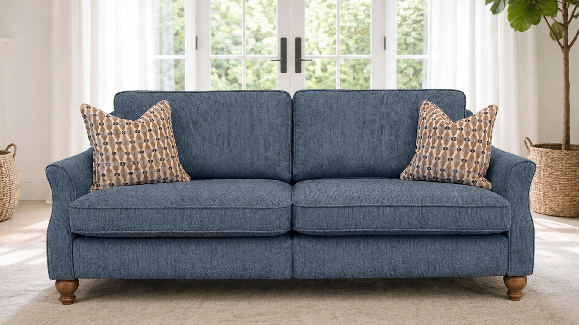 A two-seater loveseat sofa upholstered in textured blue fabric, styled with two patterned throw pillows and wooden legs, set in a bright living room with a window and potted plant in the background.