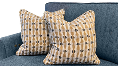 Two decorative throw pillows with a tan and cream ikat pattern featuring small black motifs, displayed on a textured blue-gray sofa.