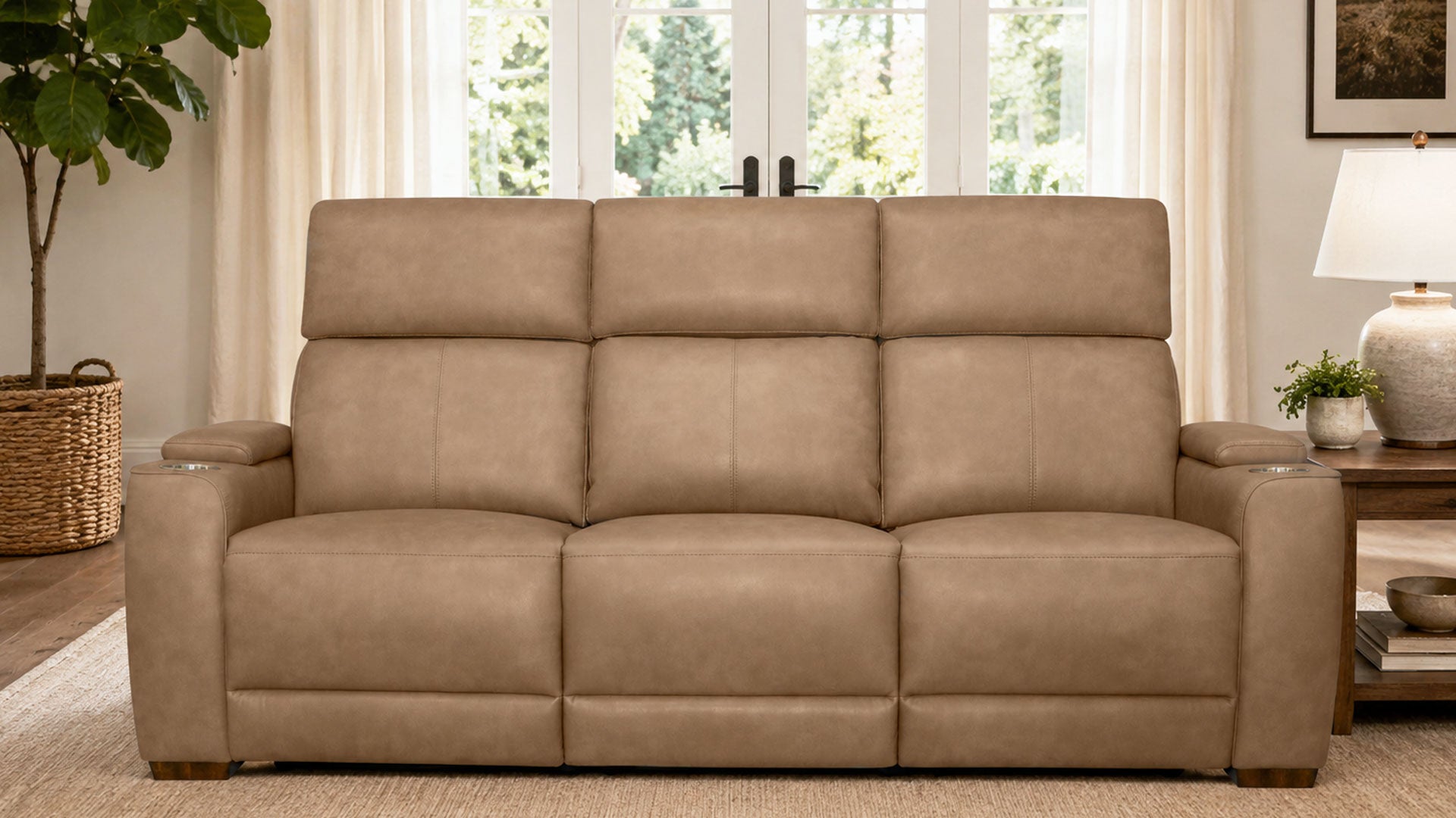 A beige upholstered reclining loveseat sofa shown in a staged living room setting with curtains, a window, a side table, lamp, and potted plant.