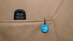 A close-up of a beige leather or faux-leather surface with a black control panel partially visible in a pocket and a small blue circular NFC tag hanging from a loop.