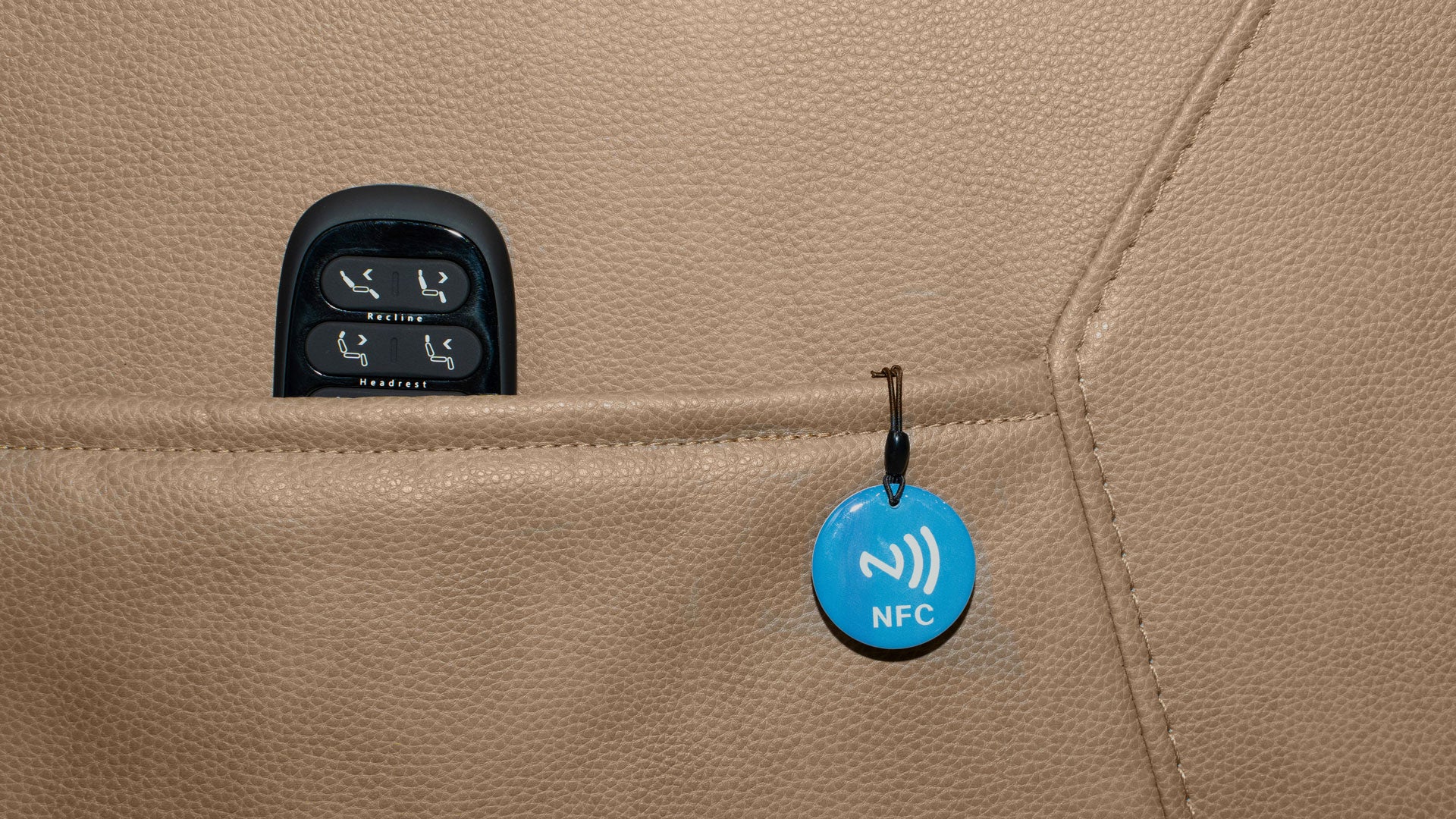 A close-up of a beige leather or faux-leather surface with a black control panel partially visible in a pocket and a small blue circular NFC tag hanging from a loop.
