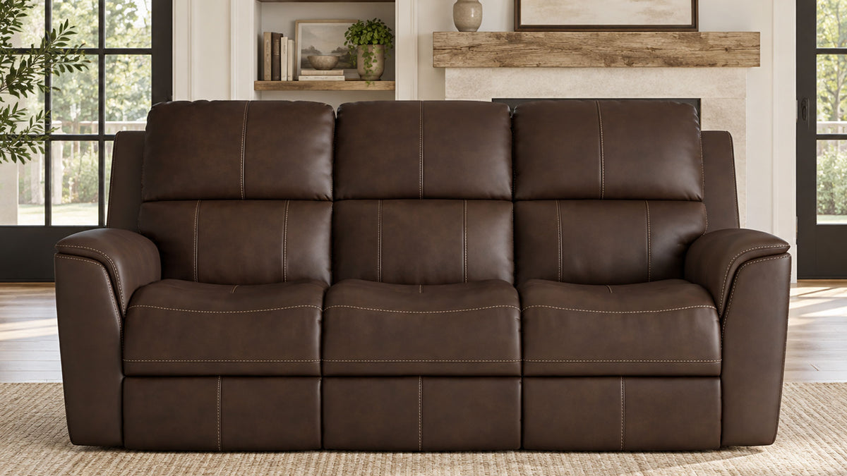 A three-seat power reclining loveseat sofa upholstered in dark brown leather, positioned in a modern living room setting with a fireplace and large windows in the background.