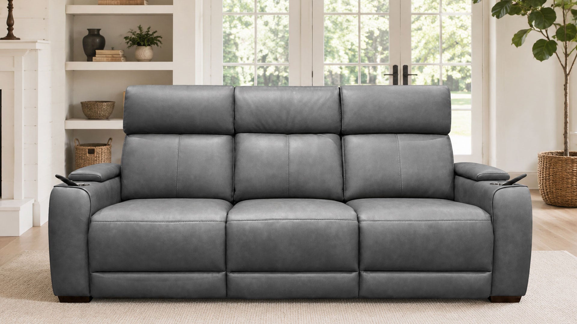 A three-seat gray leather reclining sofa positioned in a bright, modern living room with a fireplace, built-in shelves, and a potted plant.