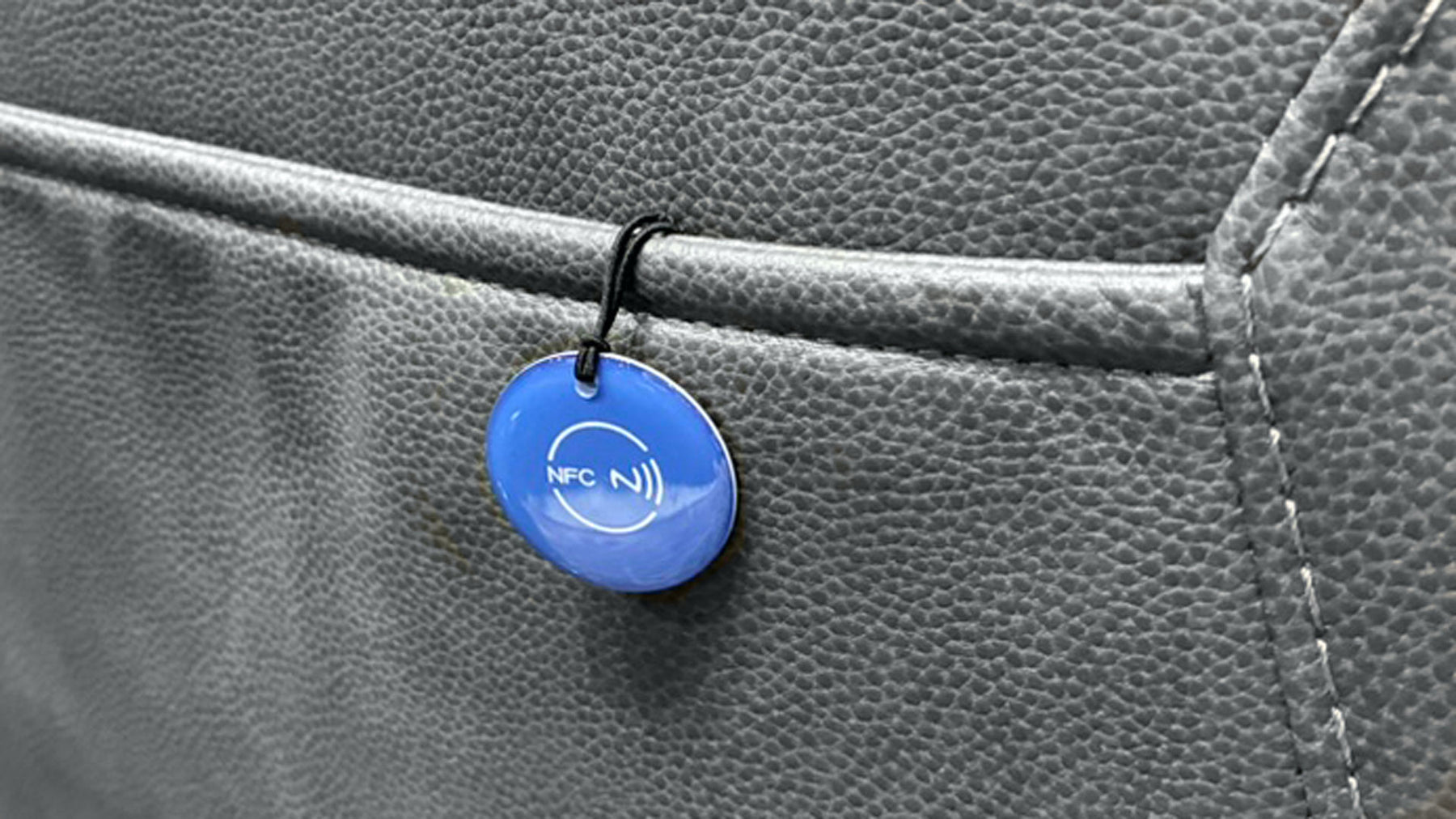 A close-up of a small round blue NFC tag on a black loop, attached to a gray textured leather surface.