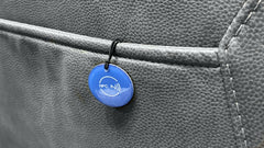 A close-up of a small round blue NFC tag on a black loop, attached to a gray textured leather surface.