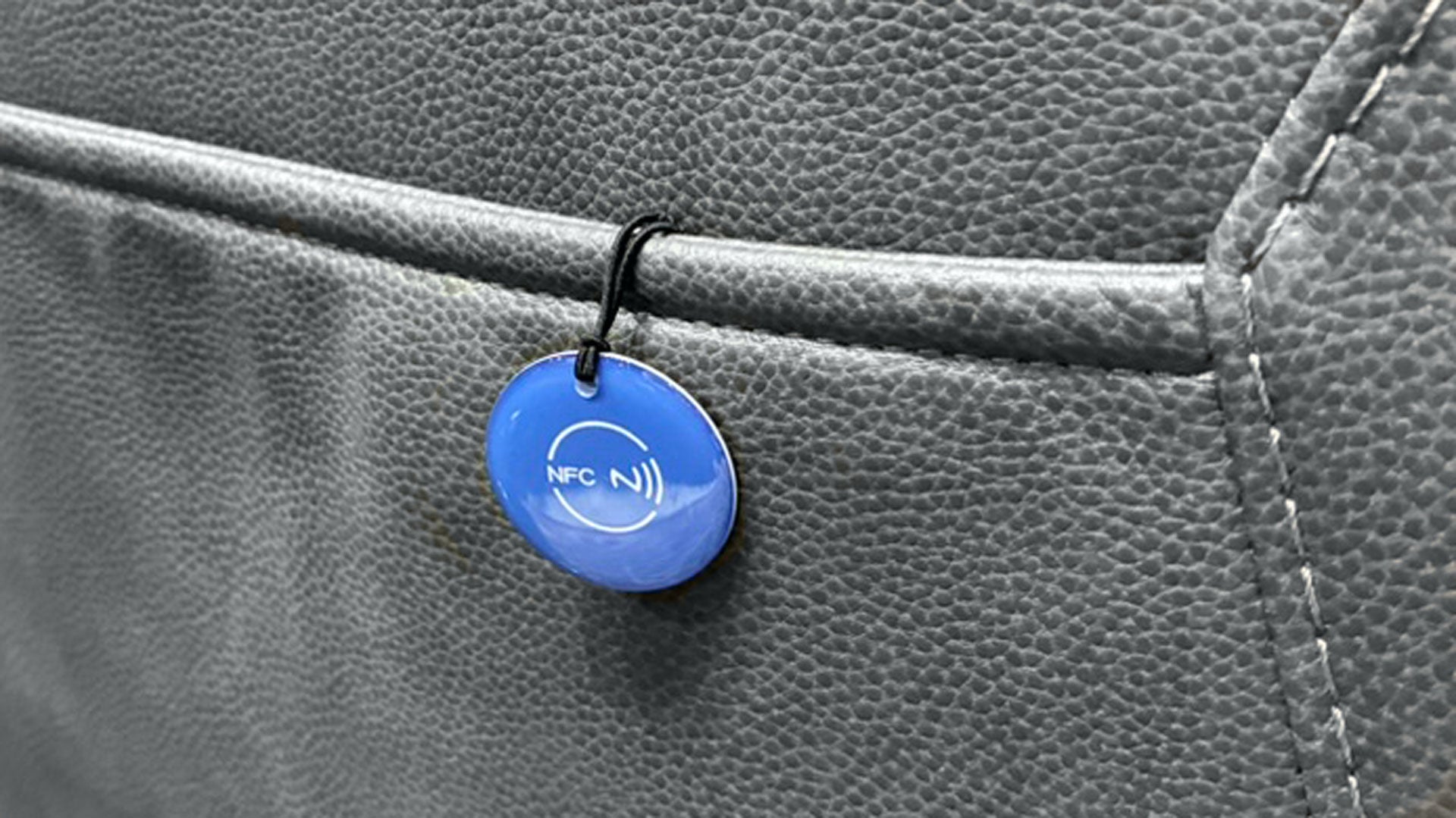 A close-up of a small round blue NFC tag on a black loop, attached to a gray textured leather surface.
