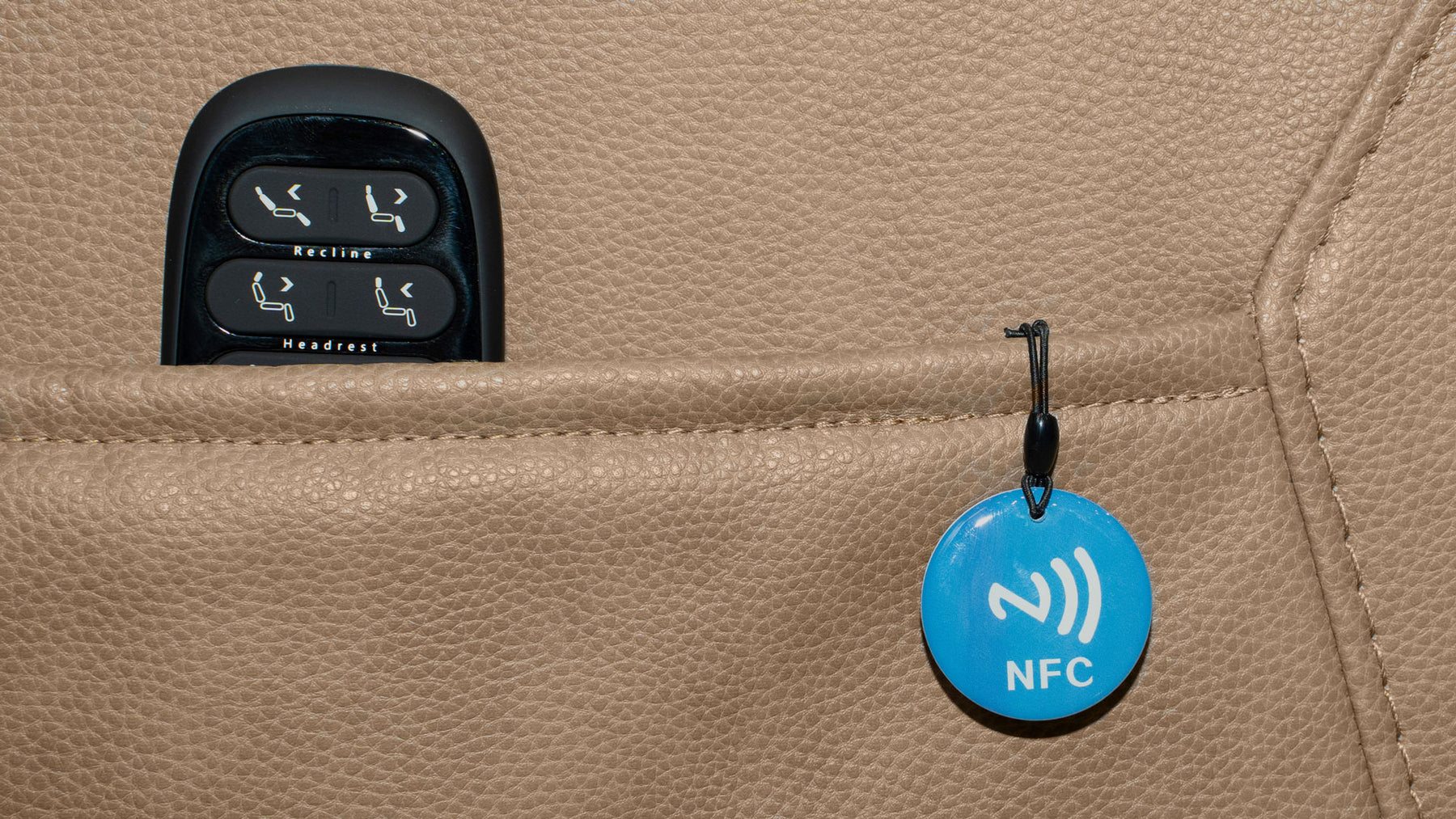 A close-up of a beige leather car seat showing a black seat control panel in a pocket and a blue circular NFC tag hanging from a black clip.
