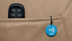 A close-up of a beige leather car seat showing a black seat control panel in a pocket and a blue circular NFC tag hanging from a black clip.