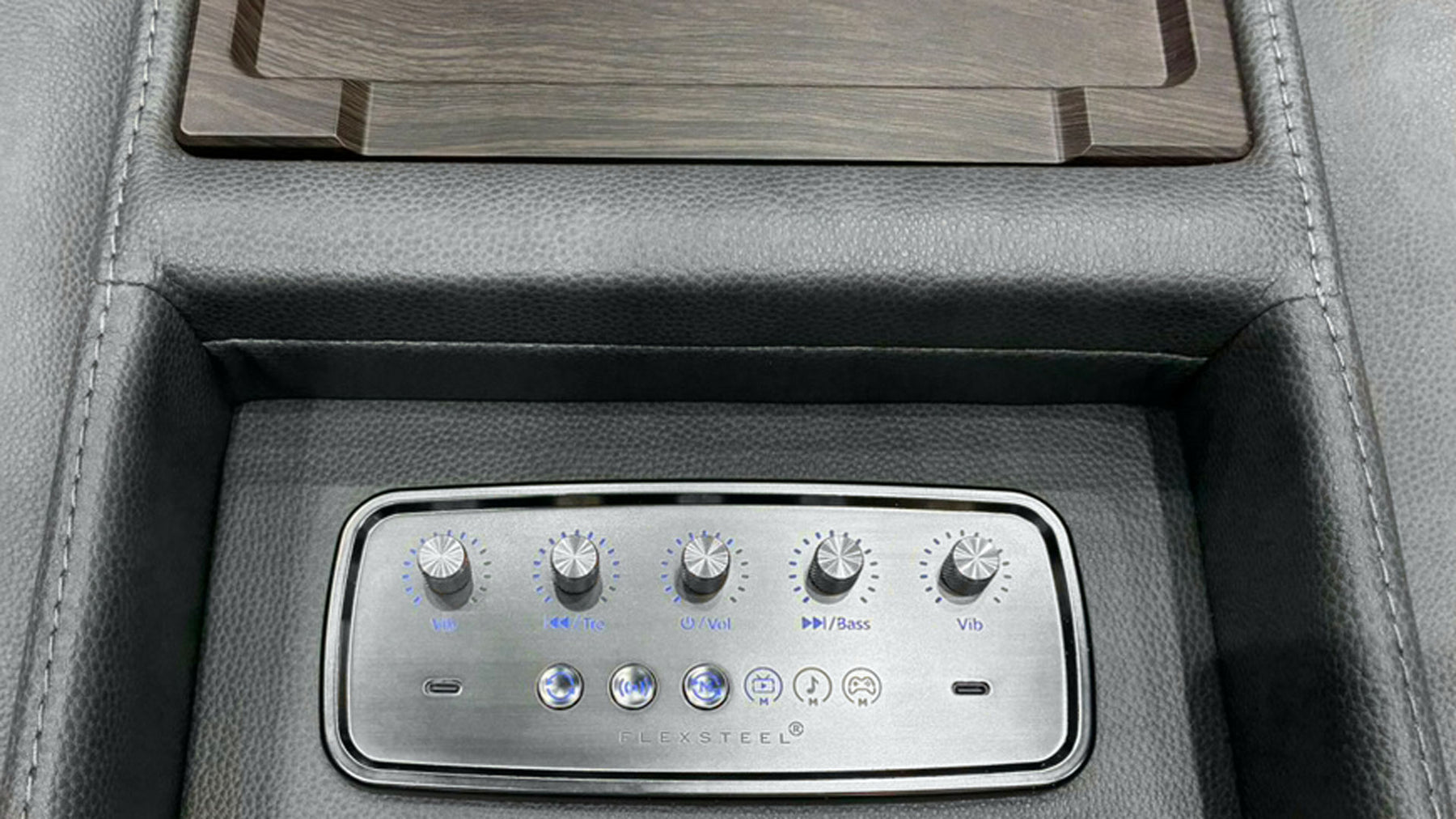 Close-up of a car center console with a silver multi-zone climate control panel set into dark gray leather, below a wood-grain tray insert.