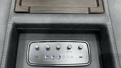 Close-up of a car center console with a silver multi-zone climate control panel set into dark gray leather, below a wood-grain tray insert.