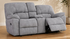 A gray two-seater recliner loveseat with a center console cup holder, shown with the right armrest reclined in a living room setting with wood floors and a potted plant.