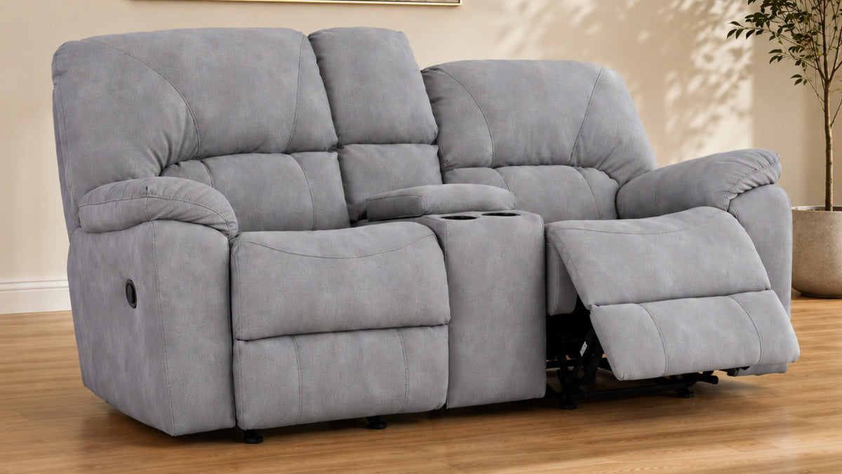 A gray two-seater recliner loveseat with a center console cup holder, shown with the right armrest reclined in a living room setting with wood floors and a potted plant.