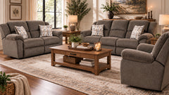 A staged living room featuring a gray upholstered recliner, loveseat, and armchair arranged around a wooden coffee table on a large patterned area rug. The room includes windows, lamps, side tables, plants, and wall art.