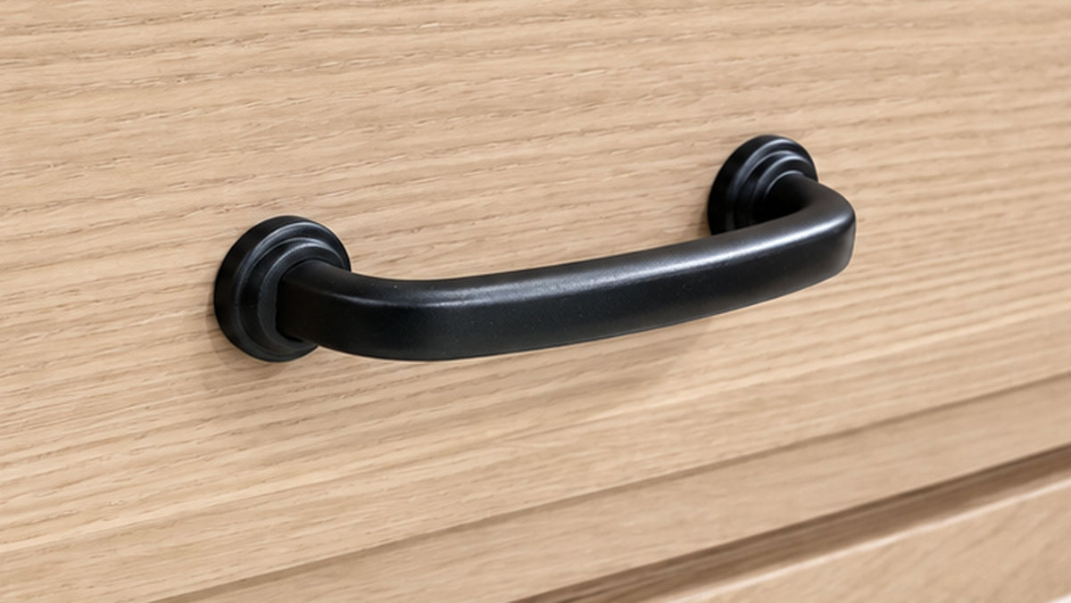 A matte black metal drawer pull handle mounted on a light oak wood cabinet door.