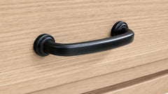A matte black metal drawer pull handle mounted on a light oak wood cabinet door.