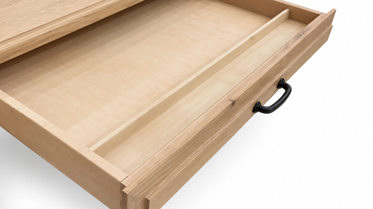 A close-up of a light natural wood drawer pulled open, showing a recessed interior divider and a black metal pull handle on a white background.