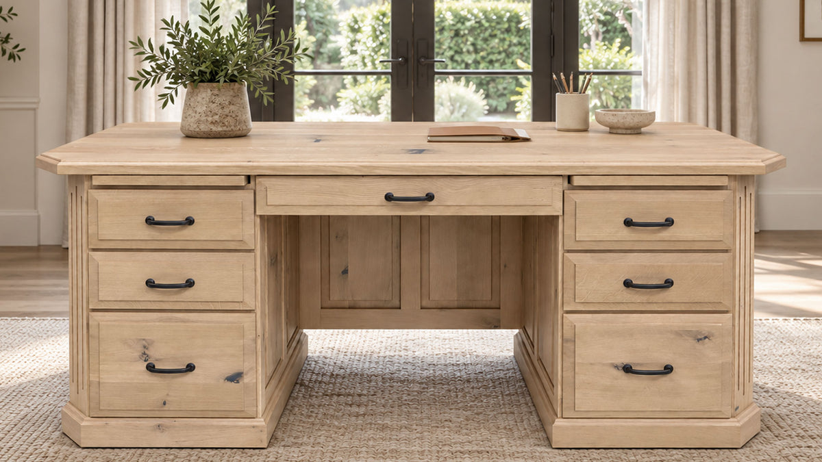 A light natural wood writing desk with six drawers and black metal handles, styled in a bright room with a potted plant and books on top.